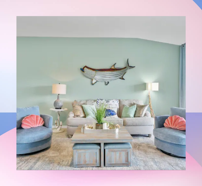 A stylish living room with light green walls and coastal decor. The space features a comfortable beige sofa with colorful throw pillows, flanked by two blue armchairs with pink shell-shaped cushions. A large wooden coffee table sits in the center, and the room is accented with two table lamps and a decorative fish wall art above the sofa. The image has a soft pink gradient border, adding a touch of elegance.