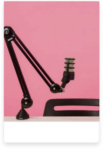 A professional microphone mounted on a black adjustable arm stand, set against a pink background. Below the microphone is the backrest of a black chair, creating a simple and focused setup for podcasting or recording.