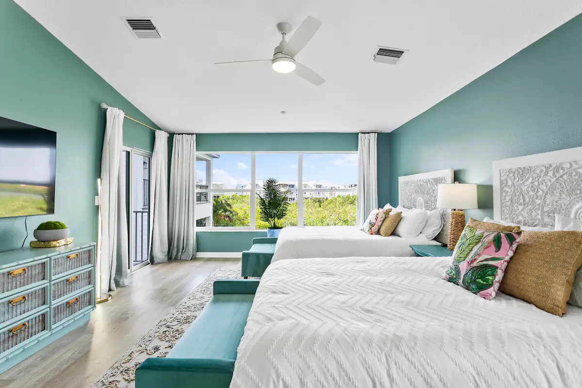 A bright and airy bedroom with teal walls, featuring two white beds with colorful throw pillows and teal benches at the foot of each bed. The room includes a large window providing a scenic outdoor view, sheer white curtains, and a ceiling fan. A flat-screen TV is mounted on the wall opposite the beds, and a stylish teal dresser with decorative accents sits below it. The decor blends modern and coastal elements, creating a fresh and inviting atmosphere.